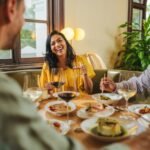 Three people laughing while dining at a restaurant table with wine and food
